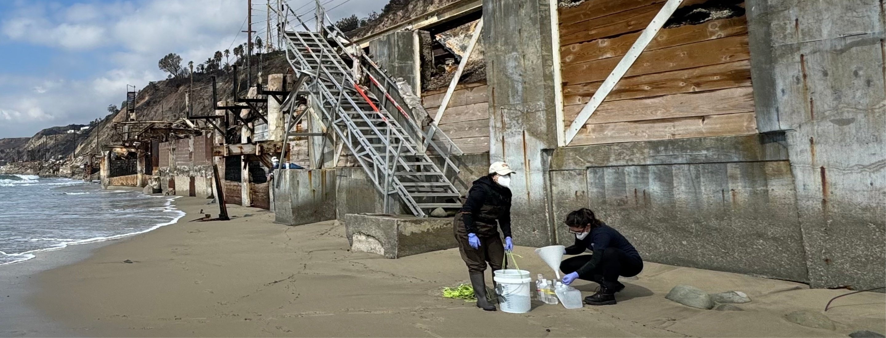 Volunteers take water samples on the sand with debris from fire in background.